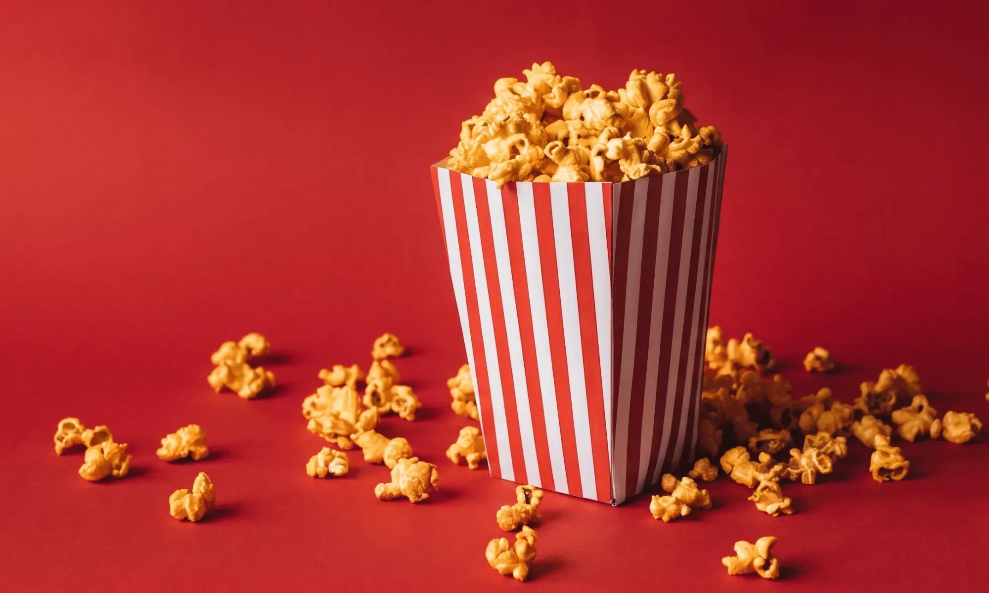 A red and white striped movie theater popcorn box overflowing with popcorn on a red background